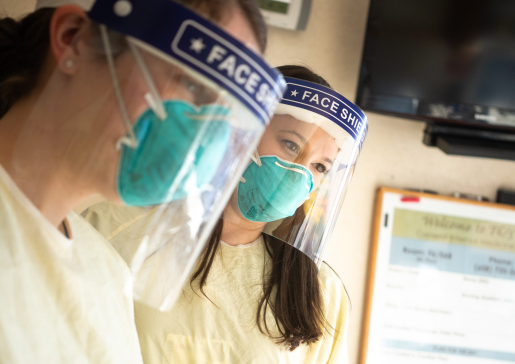 Two hospital medicine clinicians wearing masks and face shields looking down at the bed in a patient room