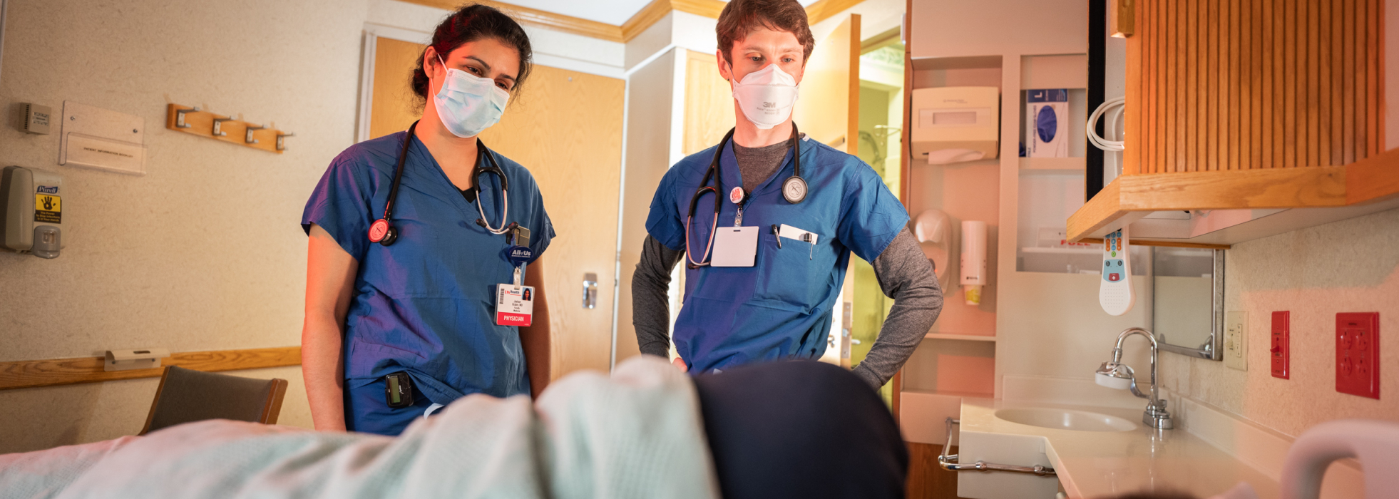 Male and female clinicians in blue scrubs and masks stand over a patient bed in the hospital