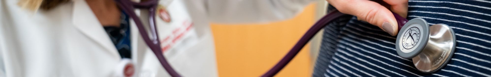 Dr. Brianna Jelenc in a white coat holds a stethoscope over the chest of a patient wearing a navy and white striped shirt