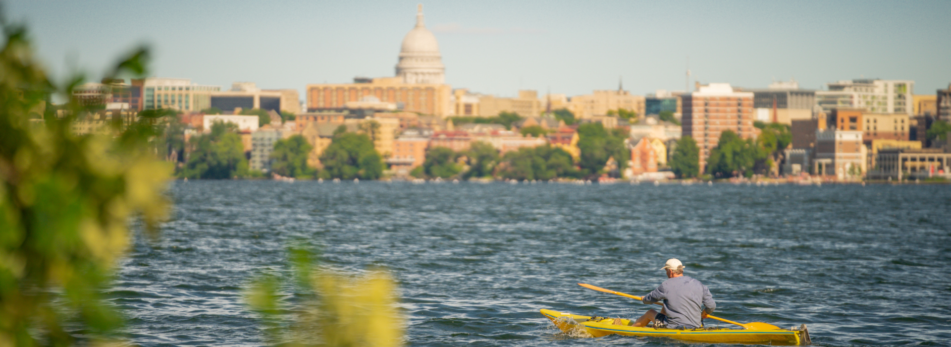 Man on kayak paddling lake Mendota with University of Wisconsin campus and Capitol building in background