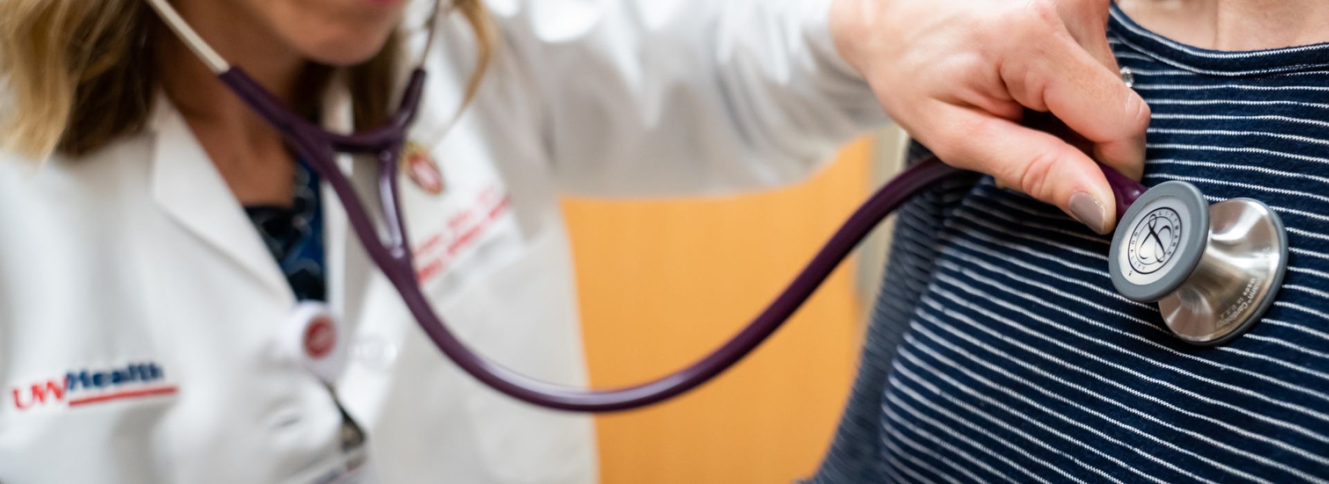 Dr. Brianna Jelenc in a white coat holds a stethoscope over the chest of a patient wearing a navy and white striped shirt