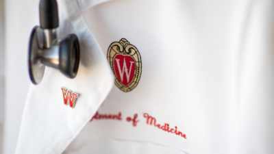 Physician&#039;s white coat embroidered with UW logo and the words Department of Medicine, with a W red enamel pin and stethoscope.
