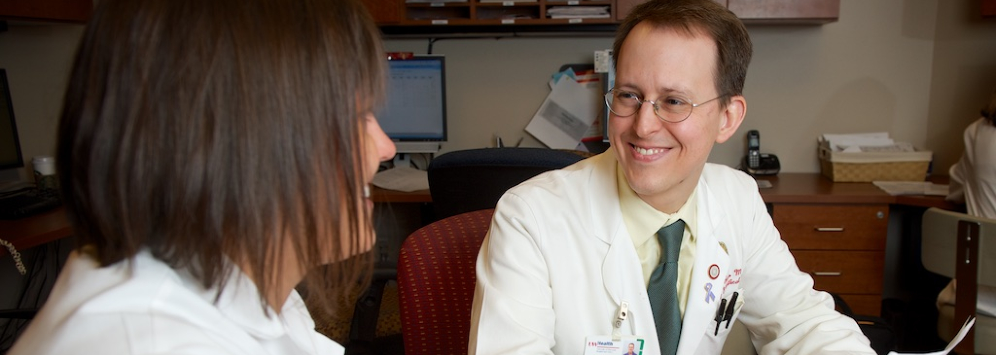 Dr. James Runo in a white coat talking with a colleague at an office table