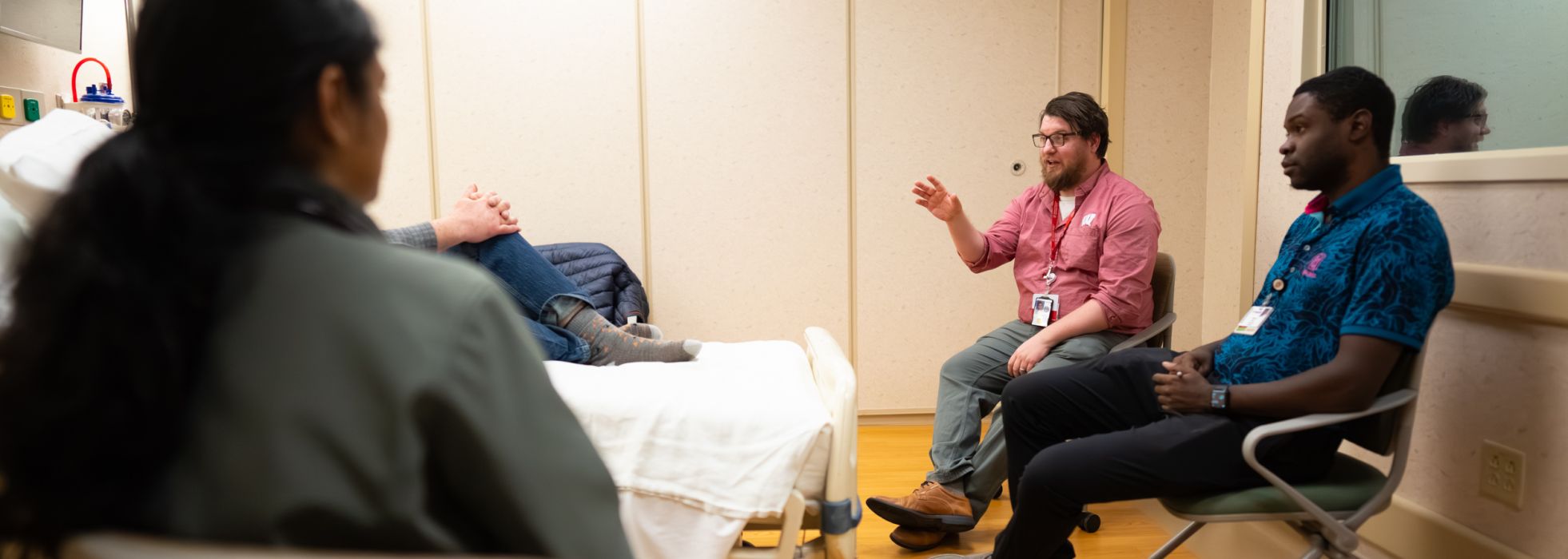 Three people sitting in a simulated patient room, with an actor sitting on the bed