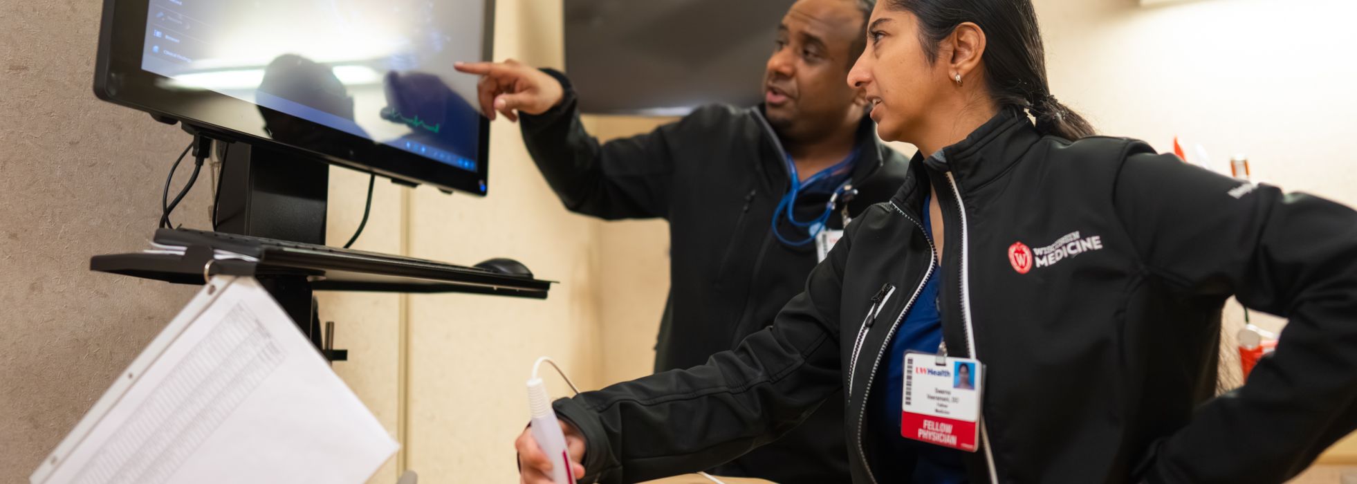 A woman performing an ultrasound on a mannequin with a man pointing at the screen