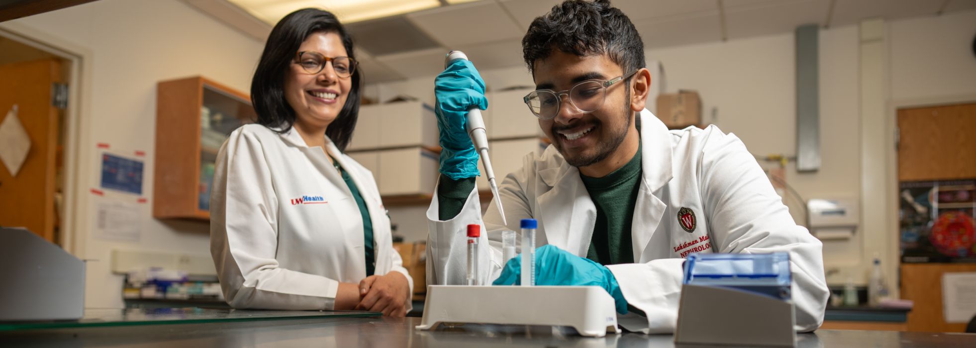 A man with a pipette and test tubes and a woman in the background supervising