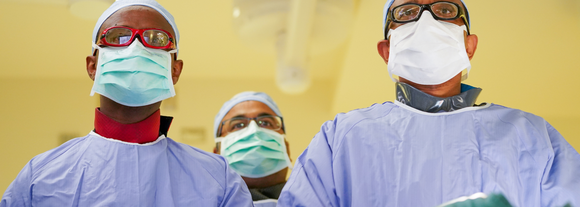 Three cardiologists in masks and gowns in the cath lab looking up at a monitor