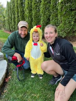 Samantha Murray-Bainer with her son James, dressed as a chicken, and her husband, Kyle, posing outside in a field with trees in the background.