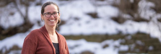 Dr. Meghan Brennan stands outside wearing dark-rimmed glasses and an orange sweater