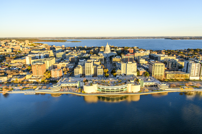 An aerial photo of Madison, with Lake Mendota in the background, the State Capitol and downtown Madison in the mid-ground, and Monona Terrace and Lake Monona in the foreground.
