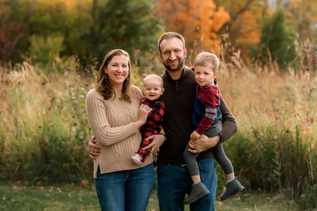 A family portrait of Dr. Murray-Bainer with husband, Kyle, and their toddler (Joshua) and baby (James) standing in a field in autumn.