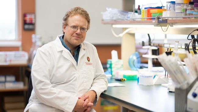 A photo of Dr. Dudley Lamming wearing a white coat and sitting at the bench in his lab.