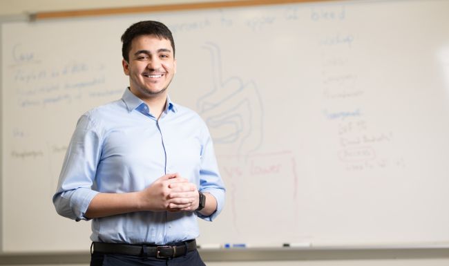Dr. Mazen Almasry standing in front of a white board in a classroom, wearing a blue button down shirt and smiling..