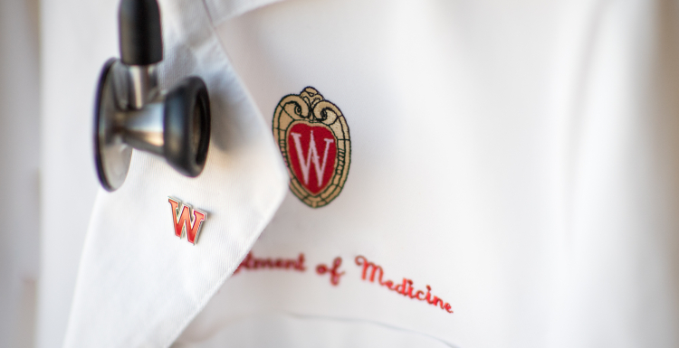 Physician&#039;s white coat embroidered with UW logo and the words Department of Medicine, with a W red enamel pin and stethoscope.