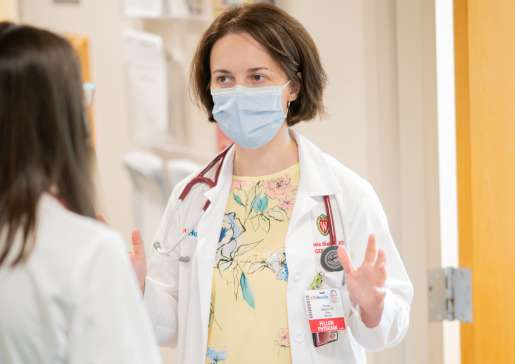 A geriatrics fellow and faculty member in white coats and masks talk in a hospital hallway
