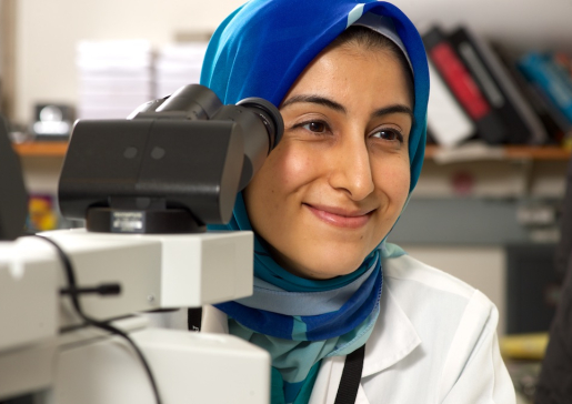 Dr. Moniba Nazeef smiles while looking out from behind a microscope