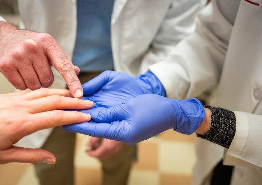 Closeup of rheumatology faculty member examining a patient's hand while a fellow points to the ring finger knuckle