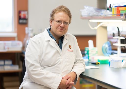 A photo of Dr. Dudley Lamming wearing a white coat and sitting at the bench in his lab.