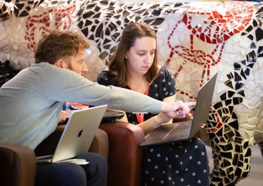 A man and woman sit together with laptops, the man pointing to something on the woman's laptop screen